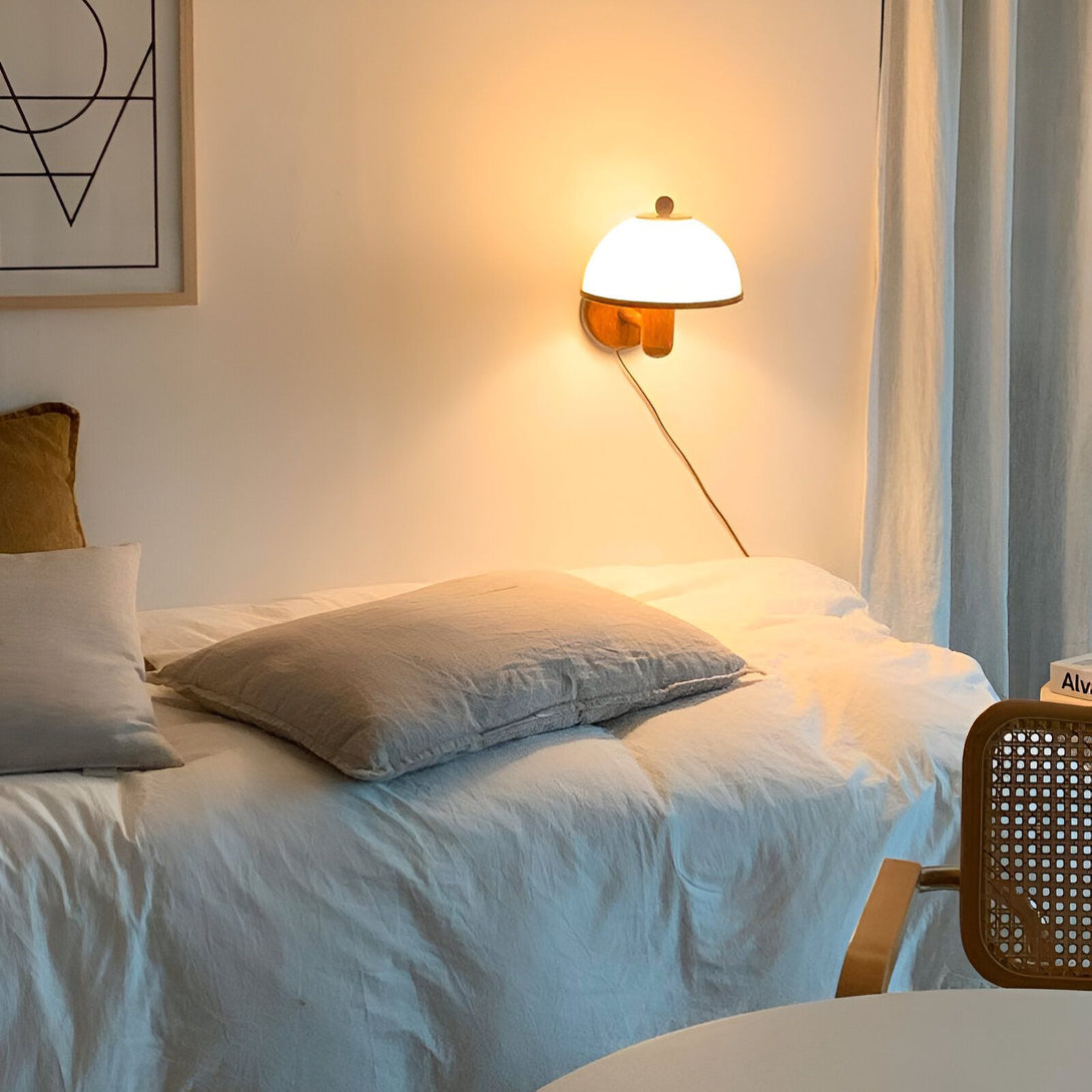Une image d'une applique murale en bois avec un abat-jour en verre blanc. L'applique est fixée au mur au-dessus d'un lit. Le lit est recouvert d'un drap blanc et de deux oreillers. Il y a une chaise en osier à côté du lit. WA-9336080733,