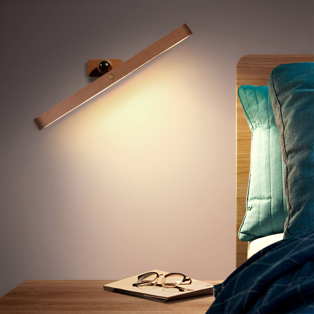 Une image d'une applique murale en bois avec une lumière chaude. L'applique est montée sur un mur beige et est allumée. Il y a une table de chevet à côté du lit avec un livre et des lunettes dessus. BE-3305924292,