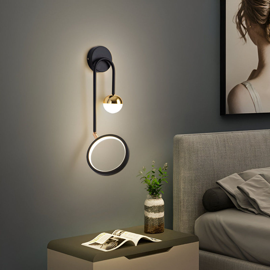 Une applique murale élégante et moderne avec une finition noire et dorée. L'applique est composée d'un cercle et d'une sphère en métal, et est fixée au mur par un support en métal noir. L'applique est équipée d'une ampoule LED intégrée et diffuse une lumière chaude et agréable. L'applique est idéale pour une utilisation dans un salon, une chambre à coucher ou un couloir.