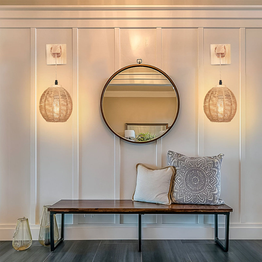 Deux appliques murales en rotin avec des ampoules allumées sont suspendues de chaque côté d'un miroir rond en bois. Un banc en bois avec un coussin à motifs est placé sous le miroir. Il y a un vase en verre sur le sol à côté du banc. ER-557575426X,