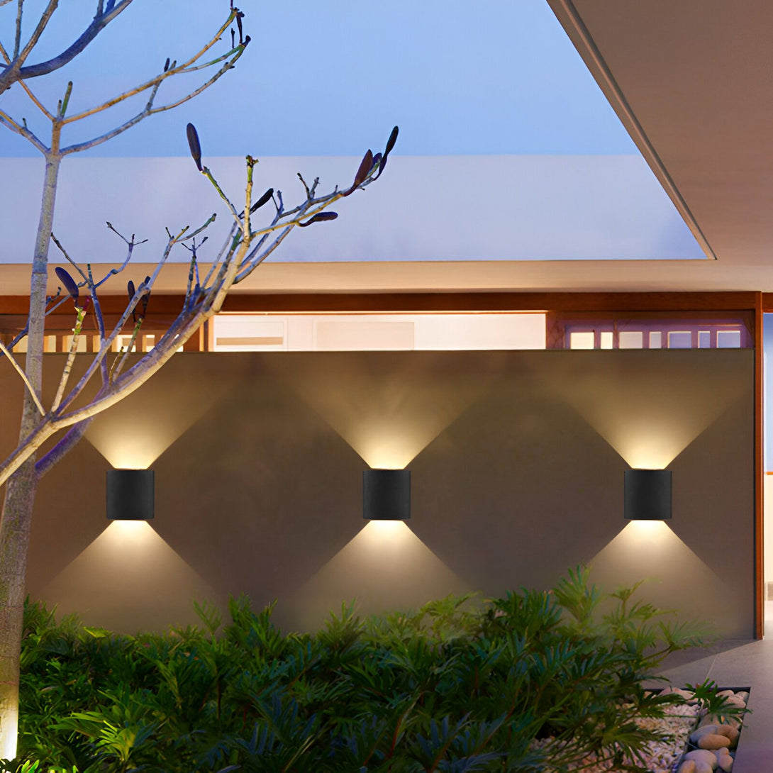 Une image d'une maison avec trois appliques murales noires. Les appliques sont en forme de cube et émettent une lumière blanche. La maison est de couleur claire et a un toit plat. Il y a des plantes vertes devant la maison et un arbre à gauche. Le ciel est bleu nuit. BO-3085302646,BO-7326999600,BO-8165967142,BO-7211273860,BO-9567856427,BO-3084783063,BO-2102062767,BO-9788980031,BO-3357184591,BO-6572328260,BO-0675009294,BO-2785354184,