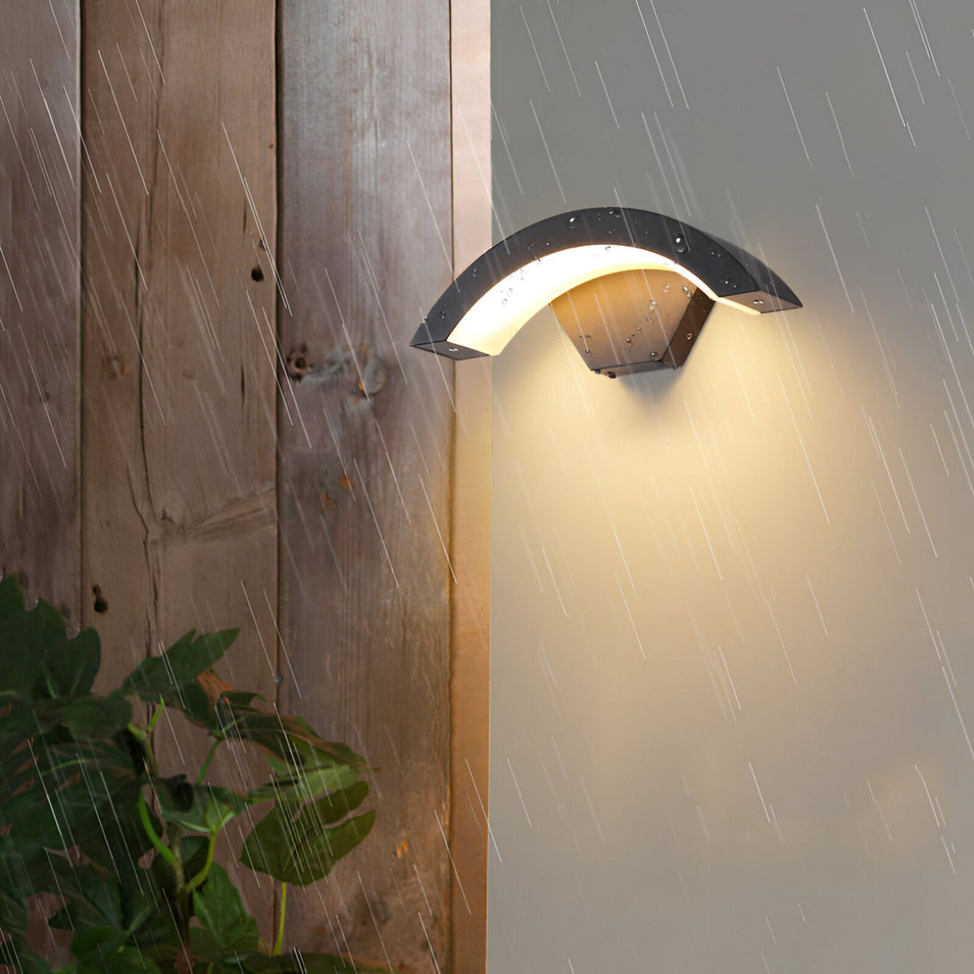 Une applique murale extérieure noire en forme de demi-lune. La lumière est allumée et on peut voir la lumière jaune qui se reflète sur le mur. Il pleut et on peut voir les gouttes d'eau sur le mur et sur l'applique. DE-3800457474,DE-0159670985,DE-7974783079,DE-3860105116,