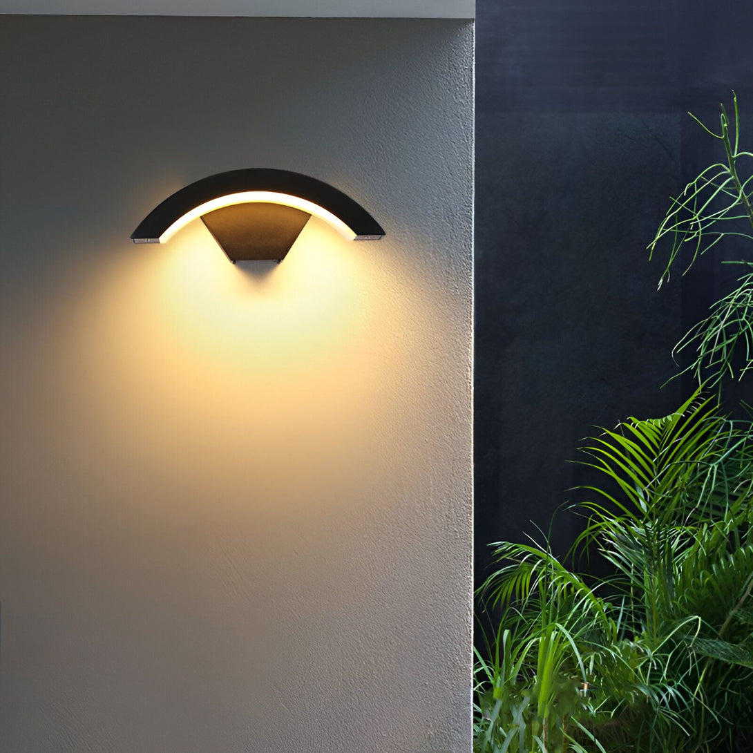 Une applique murale extérieure noire en forme de demi-lune émet une lumière chaude sur un mur beige. À côté, on peut apercevoir un feuillage vert. DE-3800457474,DE-0159670985,DE-7974783079,DE-3860105116,
