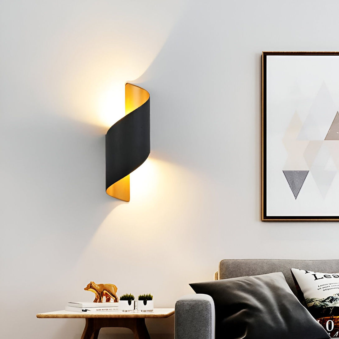 Une image d'une applique murale en métal noir et or. L'applique est en forme de spirale et émet une lumière chaude. Elle est fixée au mur au-dessus d'un canapé gris. LI-469870085X,LI-0368927849,