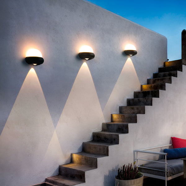 Une image d'un mur extérieur avec trois appliques murales noires. Les appliques sont en forme de demi-sphère et émettent une lumière blanche chaude. A côté de l'escalier, on peut apercevoir un pot de fleurs et un coin salon. EX-8638596960,EX-4113421196,EX-0309017424,