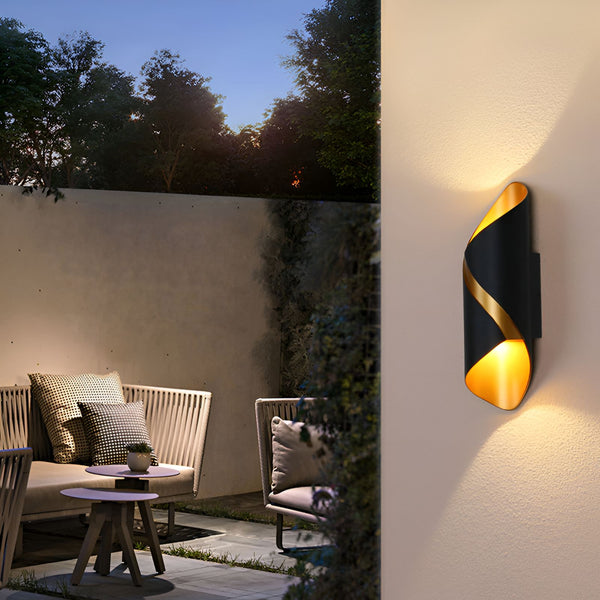 Une image d'une applique murale d'extérieur noire et dorée. L'applique est en forme de spirale et émet une lumière chaude. Elle est installée sur un mur beige, à côté d'une porte. Il y a un lampadaire et des plantes vertes dans le jardin. MA-5138903403,MA-7925922729,