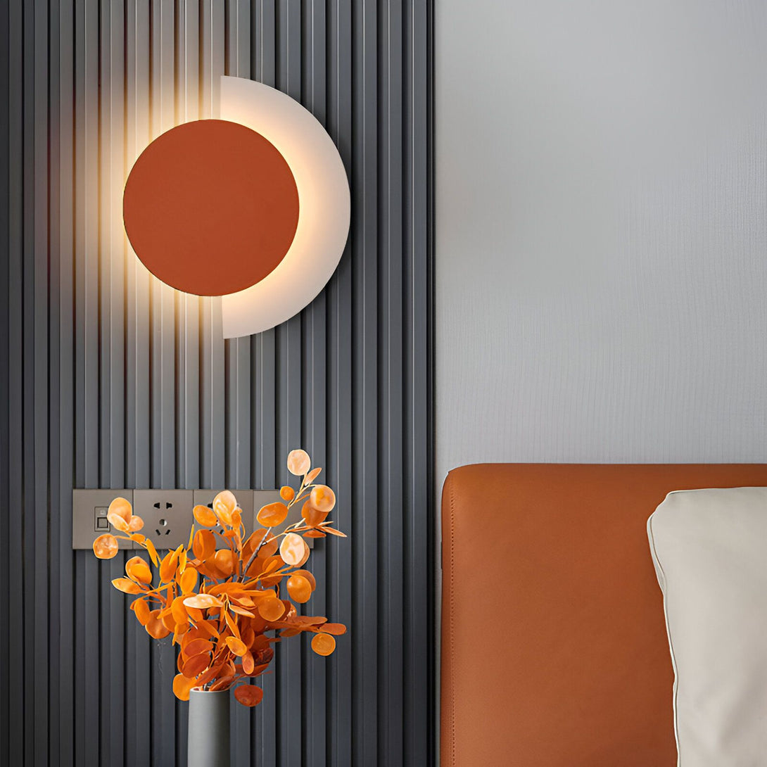 Une applique murale en forme de demi-lune orange et blanche est fixée sur un mur gris. À côté, sur une table de chevet en cuir marron, se trouve un vase contenant des fleurs séchées orangées. BE-9632631501,BE-7024548991,BE-8287115302,BE-4743076404,BE-8303465600,BE-4471555928,