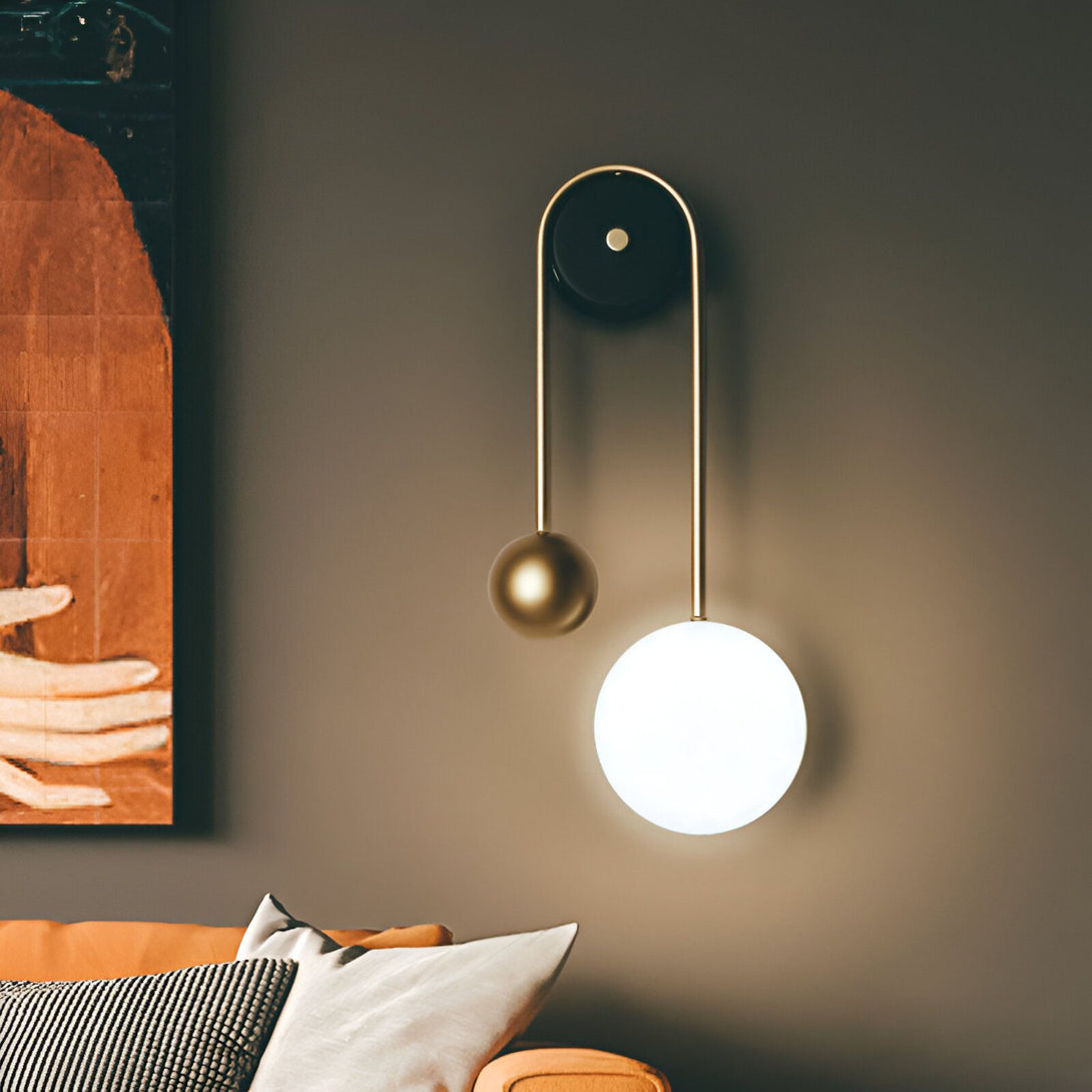 Une applique murale avec une sphère en verre blanc et une sphère en métal doré. L'applique est fixée à un mur marron foncé. À côté de l'applique, il y a un canapé orange avec des coussins blancs et gris. MI-5508556966,