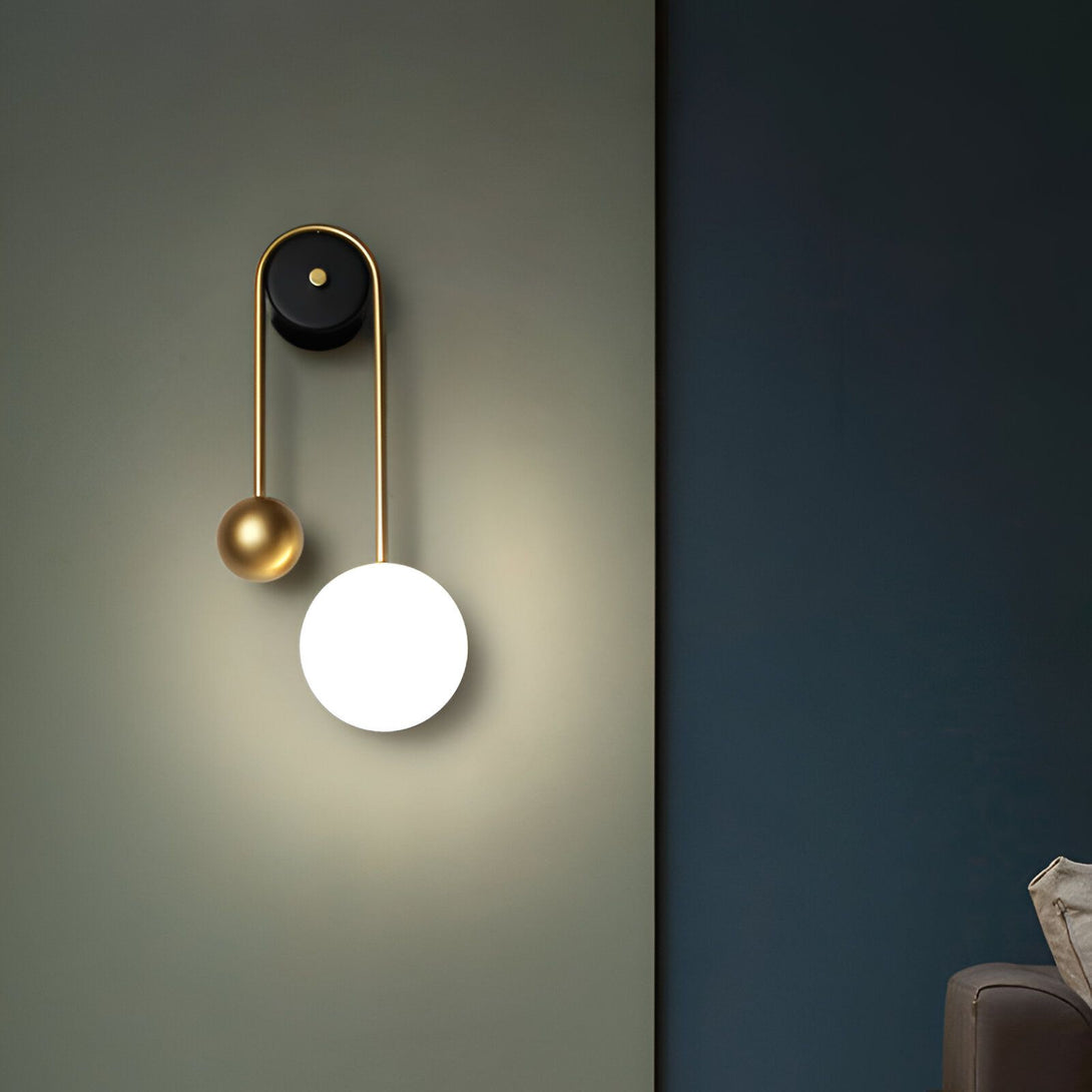 Une applique murale avec une sphère en verre blanc et une sphère en métal doré. L'applique est fixée au mur par un fil noir. MI-5508556966,