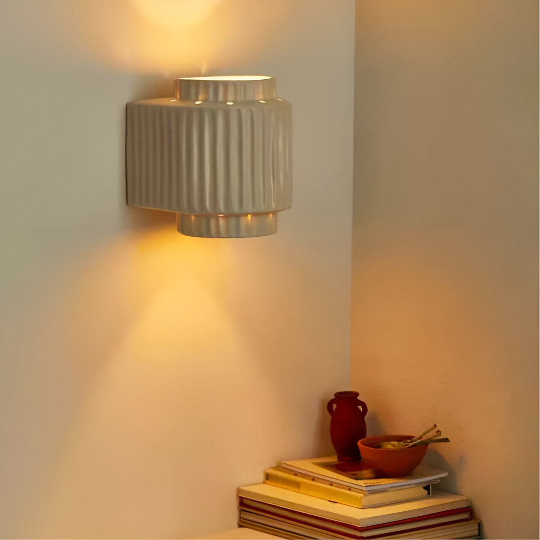 Une applique murale en céramique beige clair avec des rainures verticales. La lumière est allumée et projette une lumière chaude sur le mur. À côté de l'applique, sur une étagère, se trouvent une coupelle en céramique marron contenant des bâtonnets d'encens et un vase en céramique marron. Sous l'étagère se trouve une pile de livres et de magazines. DE-2211618871,DE-9387341801,