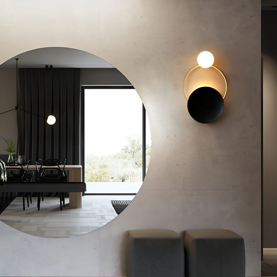 Une image d'une applique murale noire et or avec une lumière blanche et une lumière dorée. L'applique est fixée sur un mur en béton brut. À côté de l'applique, il y a un miroir rond. AL-0113600720,