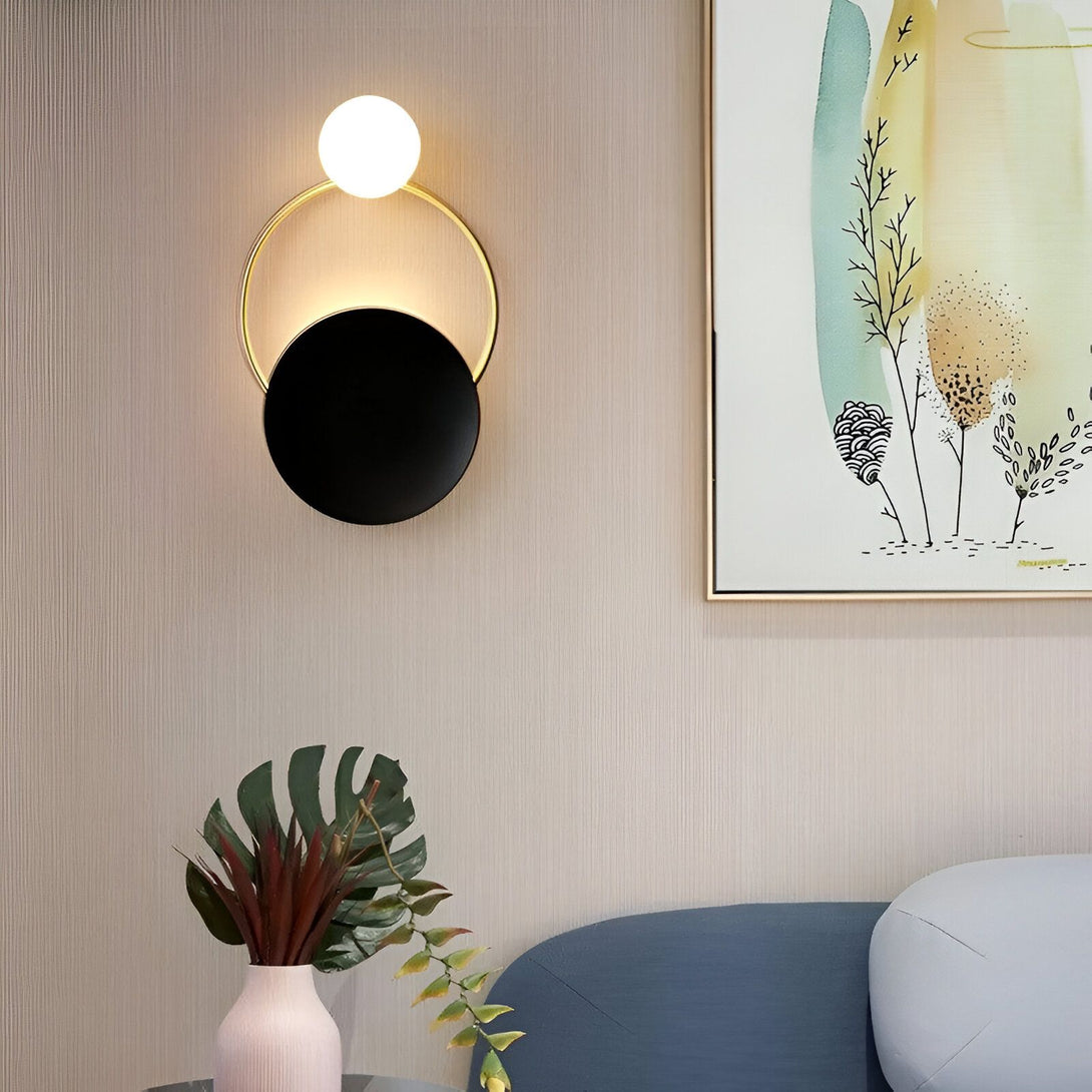 Une image d'une applique murale moderne avec un cercle doré et une sphère en verre blanc. L'applique est fixée sur un mur beige à côté d'un canapé bleu et d'une plante verte. AL-0113600720,