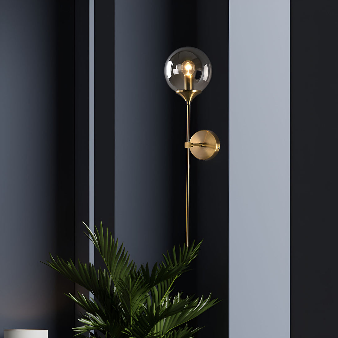 Une image d'une applique murale en laiton avec une sphère en verre transparent. L'applique est fixée sur un mur gris foncé. À côté de l'applique, il y a une plante verte. AL-5005943498,AL-6451571660,AL-2814593528,