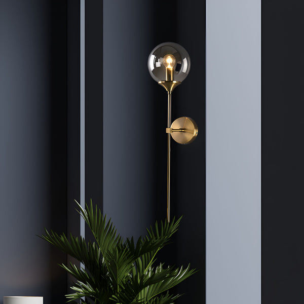 Une image d'une applique murale en laiton avec une sphère en verre transparent. L'applique est fixée sur un mur gris foncé. À côté de l'applique, il y a une plante verte. AL-5005943498,AL-6451571660,AL-2814593528,