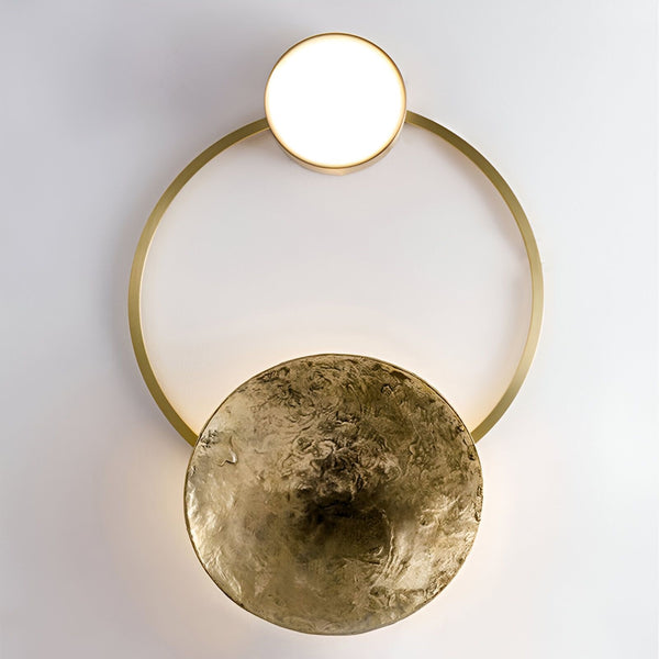 Une applique murale en cuivre antique doré et martelé. L'applique est composée d'un cercle en cuivre martelé et d'un disque en cuivre doré. Le disque est fixé au mur et le cercle est suspendu au disque. L'applique est éclairée par une ampoule LED intégrée. SO-6375224527,