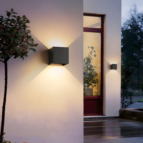 Une image d'une applique murale extérieure noire en forme de cube. La lumière est allumée et projette un faisceau lumineux vers le haut et le bas. L'applique est fixée au mur à côté d'une porte d'entrée. LE-0849619742,LE-9628653377,