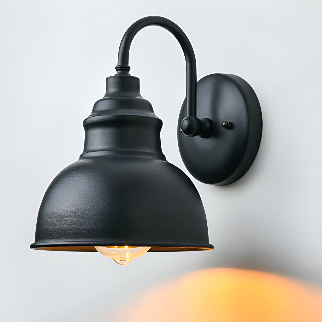Une applique murale noire avec un abat-jour en forme de cloche. L'applique est fixée au mur par une plaque ronde noire. L'abat-jour est en métal noir et a une forme de cloche. L'ampoule est visible à l'intérieur de l'abat-jour. L'applique est allumée et diffuse une lumière chaude. LA-3532870835,LA-7331102605,