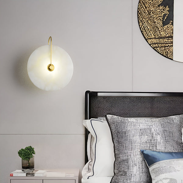 Une applique murale ronde en marbre blanc avec un bras en métal doré. L'applique est fixée au mur au-dessus d'un lit avec une tête de lit noire et des draps blancs. NA-4097404784,NA-2974006701,NA-832145366X,NA-8668627538,