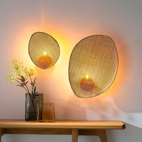 Deux appliques murales en rotin tressé, de forme ovale, avec une bougie chauffe-plat à l'intérieur. Les appliques sont fixées au mur au-dessus d'une table en bois, sur laquelle est posé un vase en verre transparent contenant des fleurs séchées. EN-7337637519,EN-7543142791,