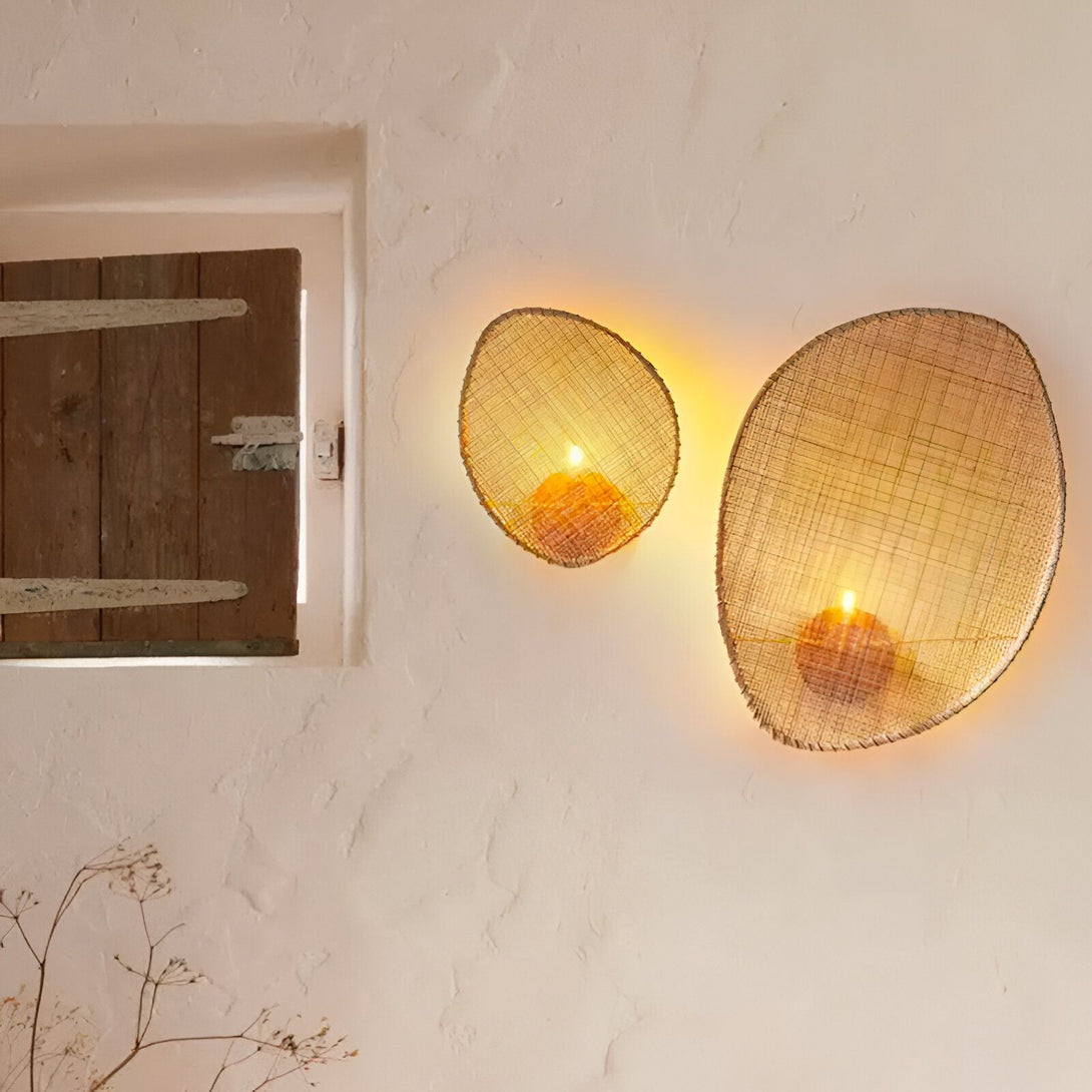 Deux appliques murales en osier tressé de forme ovale, contenant chacune une bougie allumée, sont accrochées à un mur blanc cassé. À gauche, une fenêtre en bois est entrouverte. EN-7337637519,EN-7543142791,
