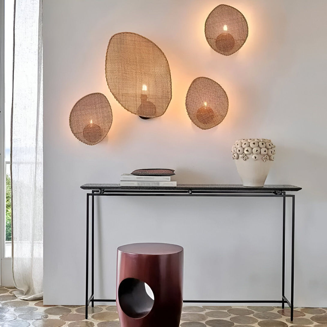 Une image d'une console en métal noir avec un vase en céramique blanche et des livres dessus. Au mur, il y a cinq appliques murales en forme de panier en osier avec une bougie allumée à l'intérieur. Un tabouret en bois avec un trou au milieu est placé devant la console. Le sol est en pierre polie. EN-7337637519,EN-7543142791,