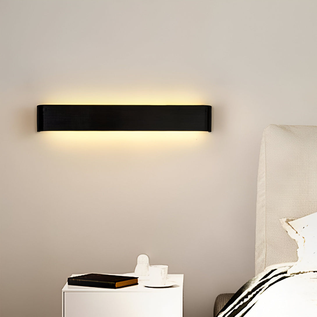 Une applique murale noire et rectangulaire émet une lumière chaude. Elle est installée au-dessus d'un lit avec une table de chevet et un livre fermé dessus. AL-1403436983,AL-0024826731,AL-5447236630,