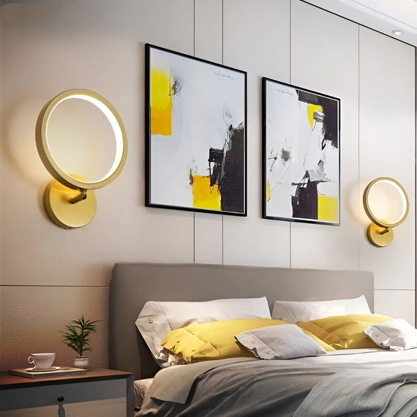 Une paire d'appliques murales rondes en métal doré avec un anneau lumineux blanc. Les appliques sont fixées de chaque côté d'un lit avec une tête de lit beige et des draps blancs et jaunes. Il y a deux tableaux abstraits accrochés au mur derrière le lit. EL-3769676734,EL-0921071280,EL-2038712905,