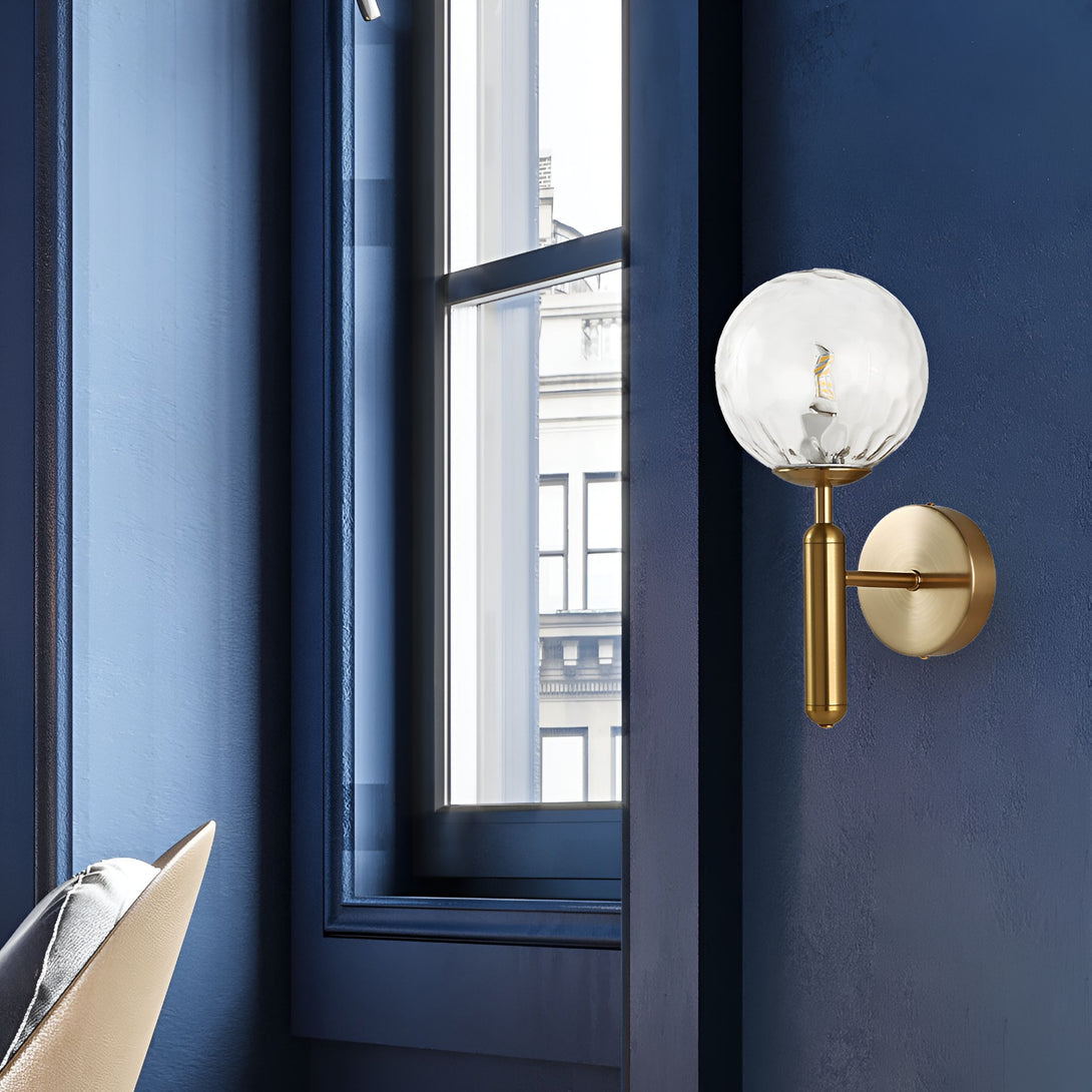 Une image d'une applique murale en laiton avec une boule en verre transparent. L'applique est fixée sur un mur bleu foncé. À côté de l'applique, il y a une fenêtre qui laisse entrer la lumière du jour. HS-6642723279,