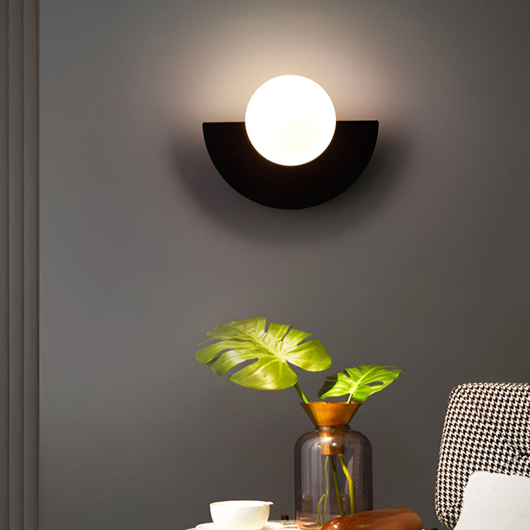 Une applique murale noire avec une forme semi-circulaire et une boule en verre opale. À côté, se trouve un vase en verre transparent avec des feuilles vertes. GU-5717952775,GU-9785984192,GU-5756525742,GU-2770483412,GU-259462313X,GU-7909977727,GU-9845973795,GU-8738437147,GU-6622229414,GU-8570558481,GU-3445094918,GU-3069247571,GU-5711512889,GU-8453761582,GU-7173945844,