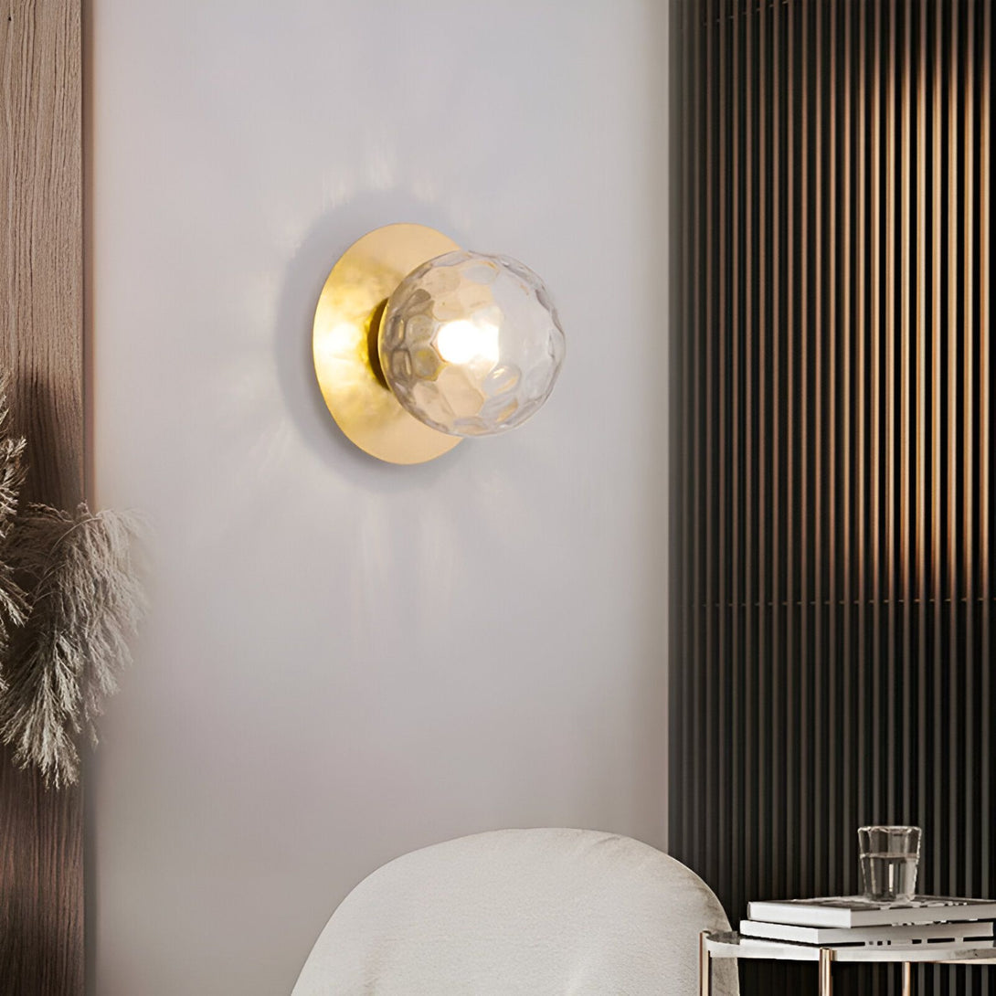 Une applique murale en forme de sphère en verre transparent avec une finition dorée. La lumière est allumée et projette des ombres sur le mur. KE-2275189483,KE-5558580682,KE-7081243789,KE-0448745380,