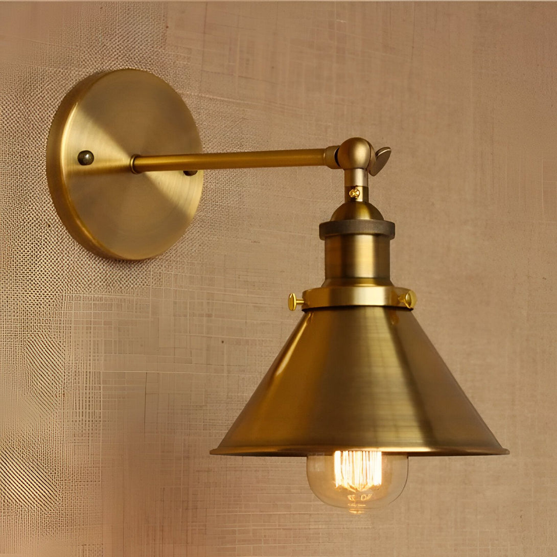 Une applique murale en laiton avec un abat-jour en forme de cône. L'applique est fixée au mur par une plaque ronde et un bras pivotant. L'abat-jour est maintenu en place par une vis. L'ampoule est visible à l'intérieur de l'abat-jour. LA-7761625263,