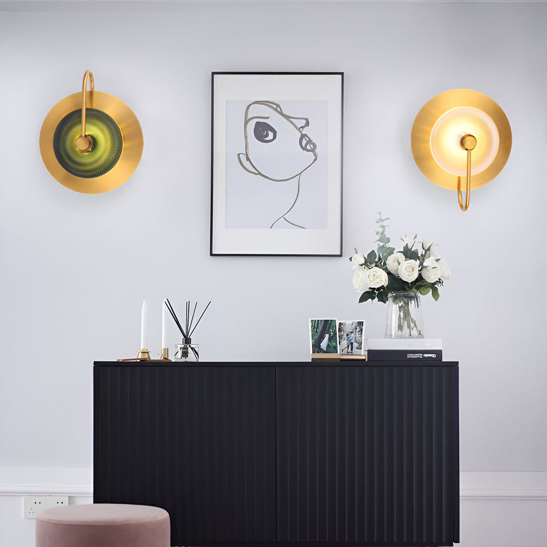 Deux appliques murales rondes en laiton avec un globe en verre vert et un intérieur doré. Les appliques sont fixées à un mur blanc au-dessus d'une console noire. Sur la console se trouve un vase en verre transparent avec des fleurs blanches, une bougie blanche, un cadre photo et un diffuseur de parfum. LE-4316598880,LE-6897461202,