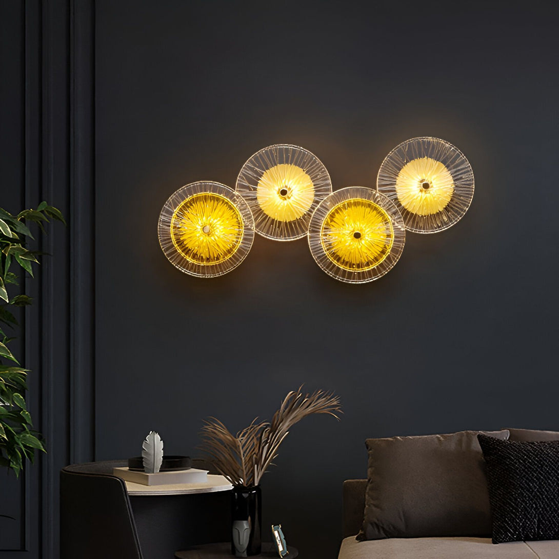 L'image représente une applique murale composée de quatre disques en verre transparent. Les disques sont disposés en ligne horizontale et sont éclairés par des LED. L'applique murale est fixée sur un mur peint en noir. AZ-1516305086,AZ-9770102490,