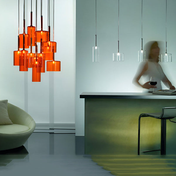 Une image de la lampe industrielle Cloche, une lampe suspendue avec plusieurs abat-jour en verre soufflé orange. La lampe est suspendue au plafond par des câbles métalliques et les ampoules sont visibles à l'intérieur des abat-jour. La lampe est allumée et émet une lumière chaude et diffuse.