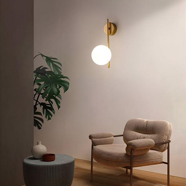 Une image d'une applique murale en laiton avec une boule en verre opale. L'applique est fixée au mur au-dessus d'un fauteuil en velours côtelé marron. À côté du fauteuil, il y a une table d'appoint avec un vase et une plante verte. GL-3736175507,GL-6321143758,GL-6843254969,GL-8040158036,GL-632193805X,GL-4001887061,GL-3434390774,GL-6755559571,