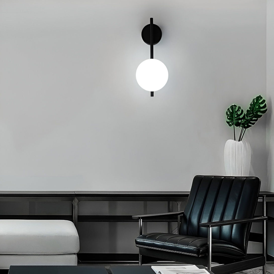 Une image d'une applique murale noire avec une boule en verre opale. L'applique est fixée sur un mur gris clair. Il y a un fauteuil en cuir noir et une plante verte à côté de l'applique. GL-3736175507,GL-6321143758,GL-6843254969,GL-8040158036,GL-632193805X,GL-4001887061,GL-3434390774,GL-6755559571,
