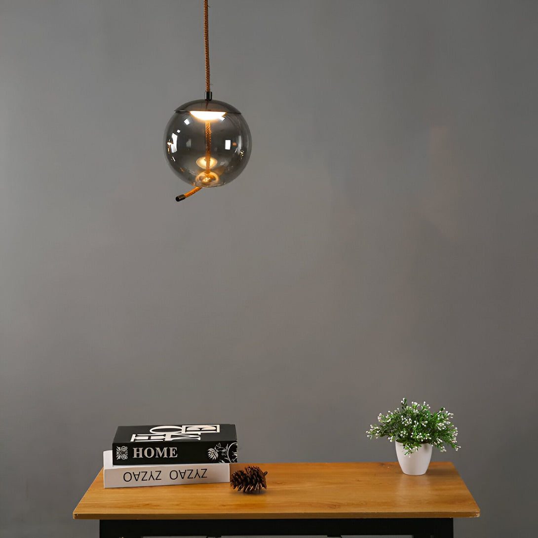 Une lampe suspendue en verre de forme sphérique de couleur gris fumé. Le luminaire est suspendu par un câble noir et est équipé d'une douille E27. La lampe est idéale pour une utilisation dans un salon, une salle à manger ou une chambre à coucher. SE-8597680784,SE-9462870446,SE-7499298493,SE-0476608740,SE-2722836270,SE-3273417943,SE-5200325382,SE-3888024714,
