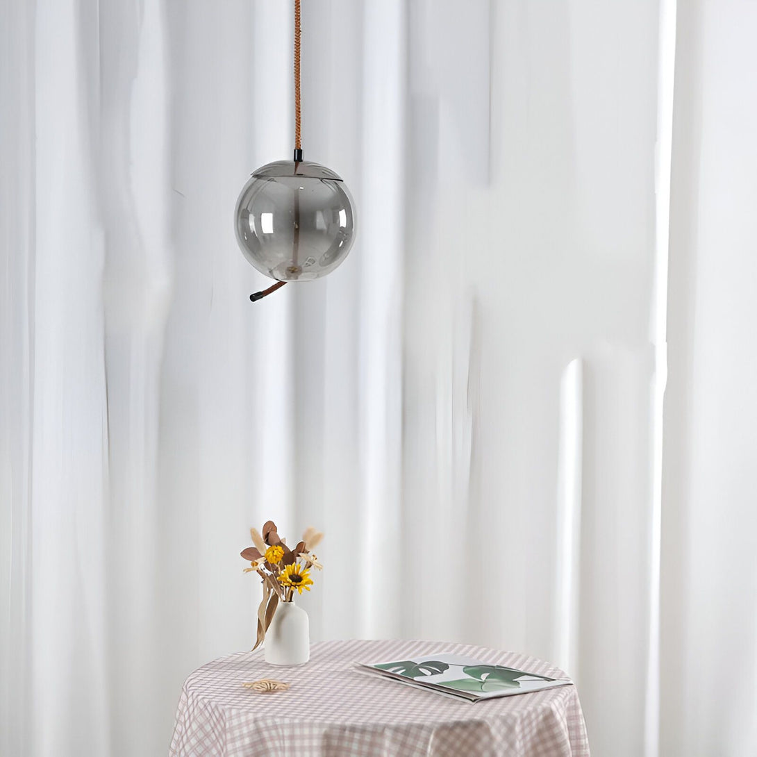 Une lampe suspendue en verre de forme sphérique est accrochée à un fil torsadé marron. La lampe est transparente avec une légère teinte fumée. Elle est suspendue au-dessus d'une table recouverte d'une nappe à carreaux blancs et roses. Sur la table se trouve un vase blanc contenant des fleurs séchées et un livre ouvert. En arrière-plan, on peut voir un rideau blanc. SE-8597680784,SE-9462870446,SE-7499298493,SE-0476608740,SE-2722836270,SE-3273417943,SE-5200325382,SE-3888024714,