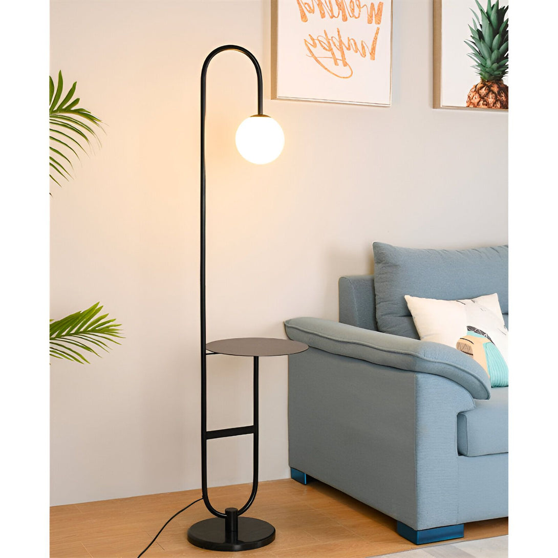 Un lampadaire noir avec une étagère ronde en métal noir. Il y a un abat-jour en verre blanc. À côté du lampadaire, il y a un canapé bleu avec des coussins blancs et bleus. LA-5213525032,LA-9607473531,LA-1687313180,
