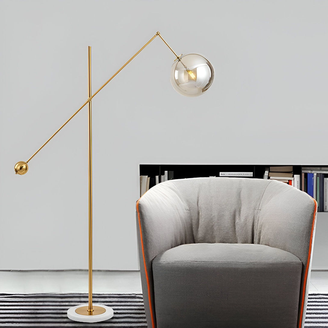 Un lampadaire doré avec une base en marbre blanc et un abat-jour en verre transparent. Il est placé à côté d'un fauteuil gris dans un salon. MA-3942036177,MA-3064936387,