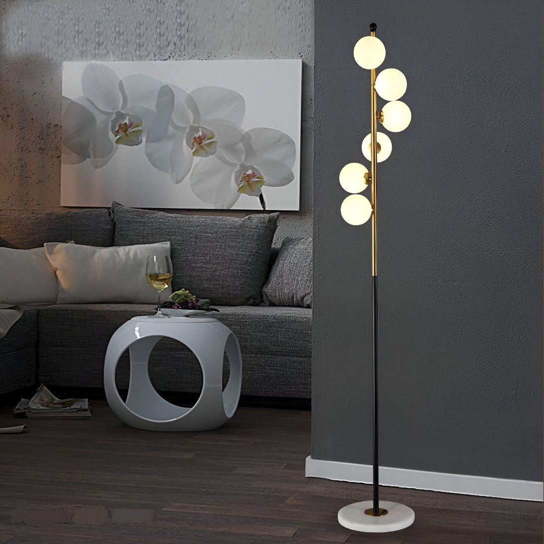 Un lampadaire noir avec cinq globes en verre blanc est placé à côté d'un canapé gris dans un salon. Il y a une table basse blanche et ronde avec un verre de vin et des raisins dessus. Un tableau de fleurs blanches est accroché au mur du fond. HS-6263847484,