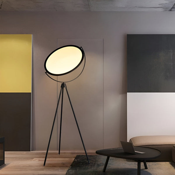Un lampadaire noir avec un abat-jour rond et blanc est posé sur un tapis rond beige dans un salon. À côté du lampadaire, il y a une table basse noire avec un ordinateur portable dessus. Le sol est en parquet clair. En fond, il y a un mur jaune et un mur gris avec un tableau noir et blanc. MA-1013807243,MA-235099841X,MA-249299418X,