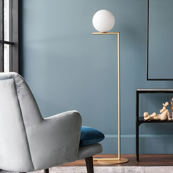 Une image d'un lampadaire doré avec une boule en verre blanc. À côté du lampadaire, il y a un fauteuil gris et une table avec un cadre dessus. Le lampadaire est allumé et diffuse une lumière douce. LS-8060965565,