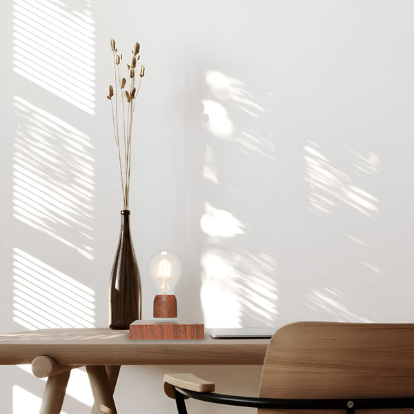 Une lampe à incandescence en lévitation au-dessus d'un socle en bois. À côté se trouve un vase en verre contenant des fleurs séchées. Le tout est posé sur un bureau en bois. En arrière-plan, on peut voir un mur blanc avec des ombres de persiennes. AL-5617346911,