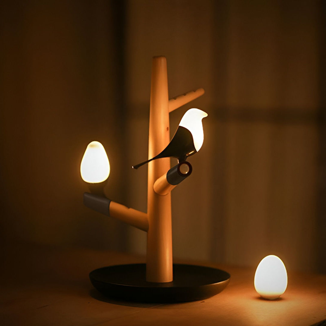 Une lampe en bois en forme d'arbre avec un oiseau perché sur une branche. L'oiseau et les feuilles sont noirs et le tronc est marron. La lampe est posée sur une table et est allumée. BI-6459513236,BI-828669189X,
