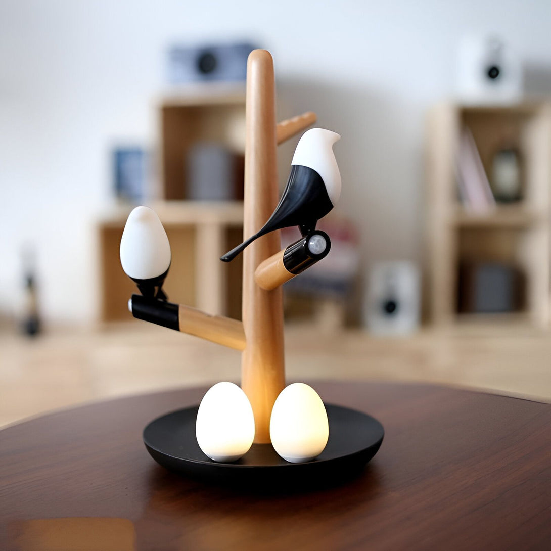 Une lampe en bois en forme d'arbre avec deux oiseaux et deux œufs. Les oiseaux et les œufs sont blancs et noirs. La lampe est posée sur une table en bois. BI-6459513236,BI-828669189X,