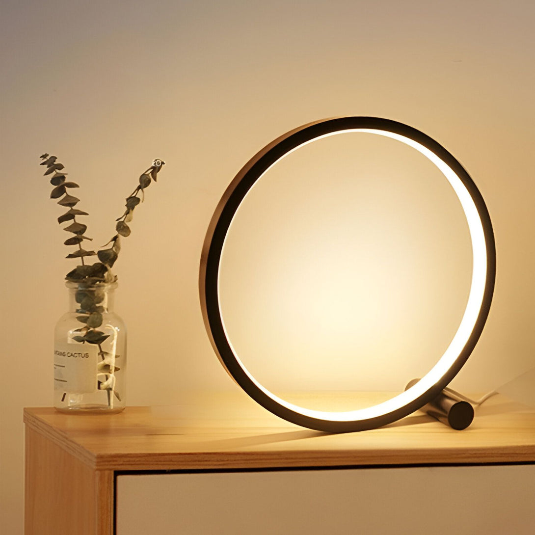 Une lampe de table LED noire en forme d'anneau avec une lumière blanche brillante. À côté d'elle, sur une table en bois, se trouve un vase en verre transparent contenant une plante verte. CE-4626626394,CE-0944412238,CE-2511648865,CE-0068266804,