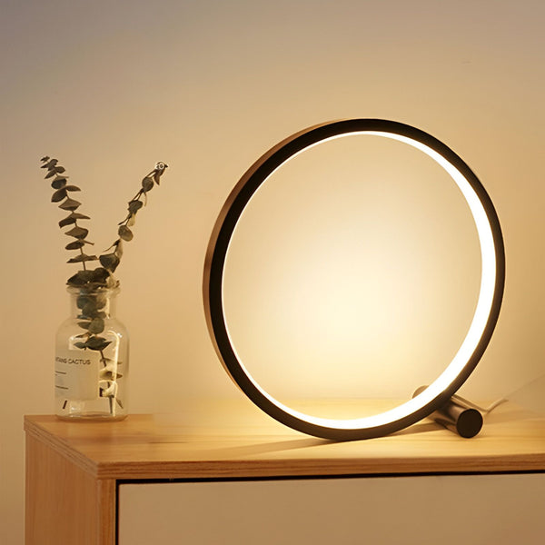 Une lampe de table LED noire en forme d'anneau avec une lumière blanche brillante. À côté d'elle, sur une table en bois, se trouve un vase en verre transparent contenant une plante verte. CE-4626626394,CE-0944412238,CE-2511648865,CE-0068266804,