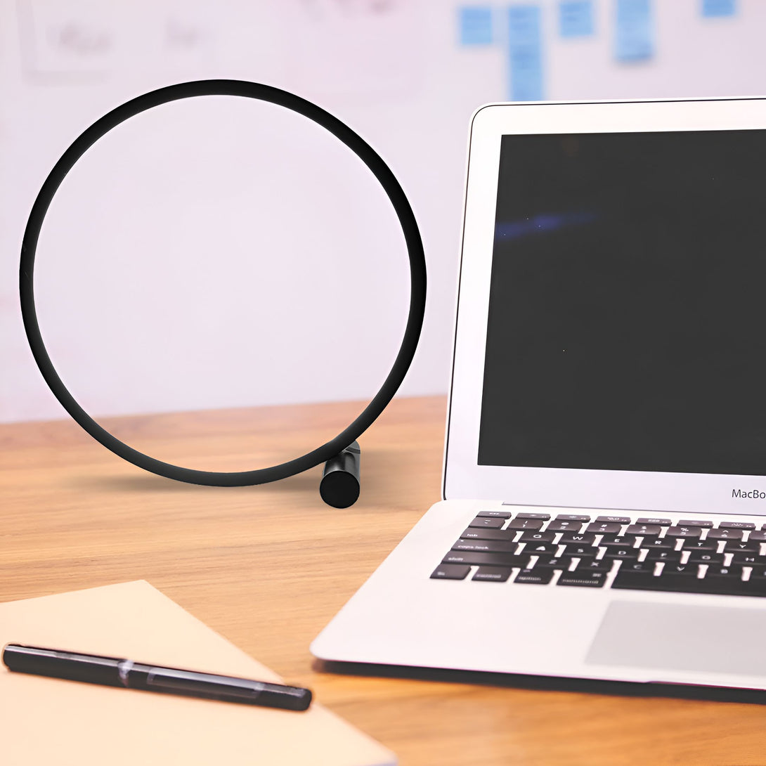 Une lampe noire en forme de cercle est posée sur un bureau en bois. À côté d'elle, se trouve un ordinateur portable MacBook ouvert. Sur le coin du bureau, à gauche, un stylo noir est posé sur un carnet de notes. CE-4626626394,CE-0944412238,CE-2511648865,CE-0068266804,