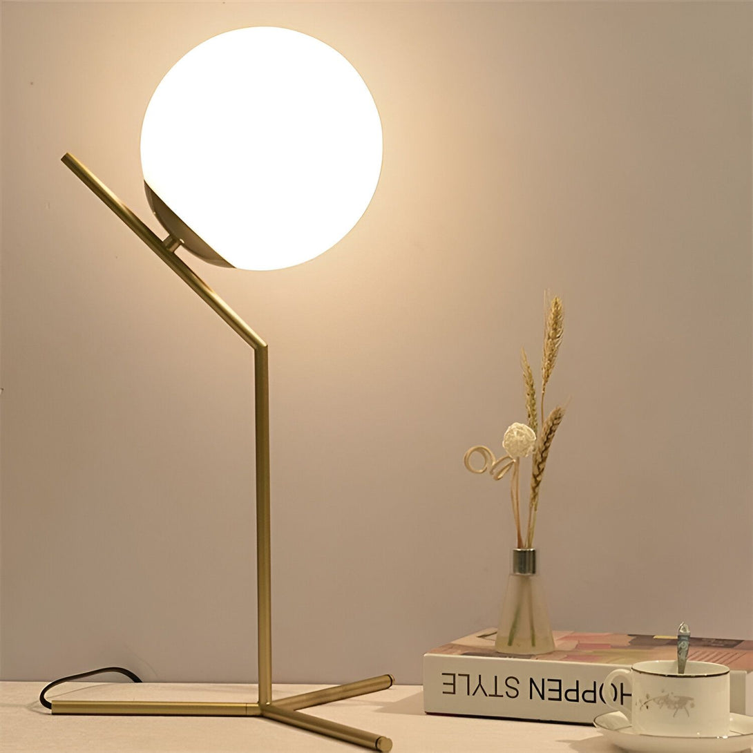 Une lampe de table moderne avec une base en métal doré et un abat-jour en verre blanc. La lampe est posée sur une table à côté d'un vase et d'un livre. CA-2757427814,CA-2757537830,